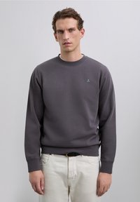 Un bărbat cu păr scurt închis la culoare poartă un hanorac gri închis cu un mic logo cu un craniu și pantaloni off-white, stând pe un fundal simplu.