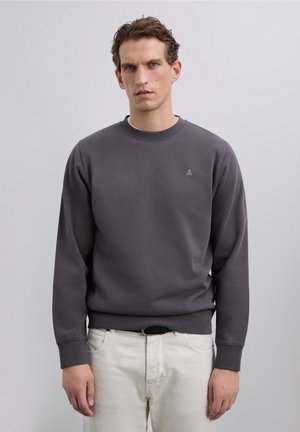 Man met kort bruin haar draagt een donkergrijze sweatshirt met een klein schedel logo en gebroken witte broek, staand tegen een effen achtergrond.
