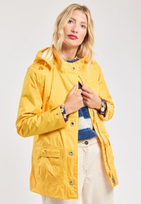Veste de pluie jaune avec fermeture éclair et boutons-pression, capuche réglable et poches avant. L'intérieur présente un pull bleu à motif.