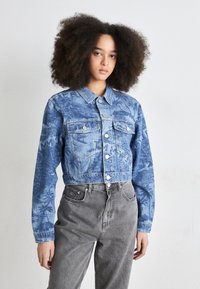 Cropped denim jacket in light blue with palm tree print, featuring a collared neck, button closure, and two chest pockets. Gray pants worn below.