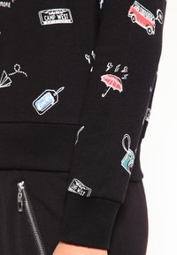 Sweatshirt noir avec des motifs brodés colorés, comprenant un appareil photo, un parapluie et divers étiquettes sur le thème du voyage sur les manches et le corps.