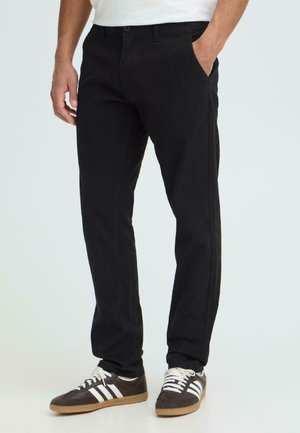 Homme portant un pantalon droit noir et des baskets noires à rayures blanches, debout avec une main dans la poche, sur fond uni.