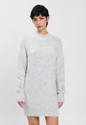 Vrouw met kort donker haar, gekleed in een lichtgrijze, zachte trui-jurk met subtiele paillettenpatronen op een effen achtergrond.