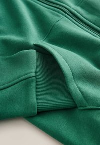 Sweatshirt zippé vert avec un tissu lisse, des poignets côtelés et une finition soignée. Comprend une seule fermeture éclair et des détails de couture minimes.