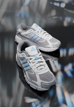Par de zapatillas Adidas blancas y azul claro para correr con partes superiores de malla y sintéticas descansando sobre una superficie reflectante.