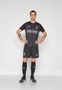 Camiseta de fútbol negra con un patrón de malla, logo del patrocinador blanco, pantalones cortos grises, calcetines negros y botines negros, con la marca Puma.