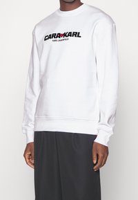 Sudadera de algodón blanca con cuello redondo, puños acanalados y un atrevido estampado gráfico en negro y rojo "GARA♥KARL" en la parte frontal.