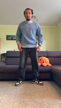 Helles blaues geripptes Sweatshirt, dunkle Jeans, schwarz-weiße Sneakers mit strukturiertem Profil. Oranger Plüsch-Oktopus auf einer grauen Couch im Hintergrund.