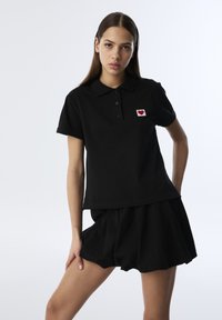 Polo negro hecho de algodón con cuello redondeado, presenta un pequeño parche en forma de corazón rojo en el lado izquierdo del pecho. Se lleva con una falda a juego.