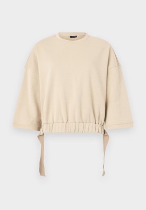 Beige avkortet sweatshirt laget av mykt materiale. Har vide ermer, en samlet midje med snorer, og rund hals.