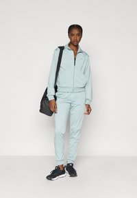 Αθλητικό μπουφάν σε ανοιχτό μπλε χρώμα με ταιριαστό παντελόνι και μαύρα αθλητικά παπούτσια· διαθέτει φερμουάρ μπροστά και ριμπ μανσέτες· συνοδεύεται από μαύρη τσάντα.