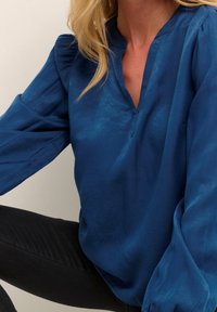 Blouse bleue en tissu soyeux avec un col en V, manches longues légèrement bouffantes et texture subtile. Présente une coupe décontractée et des détails minimalistes.