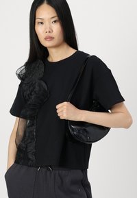 Melnas īsām piedurknēm tops ar teksturētu melnu mežģīņu volānu kreisajā pusē, apvienots ar mazu melnu ādas crossbody somu.