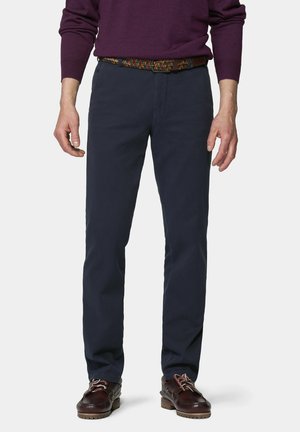 Mann trägt dunkelblaue Hose, braune Lederschuhe mit Schnürsenkeln, bunten gewebten Gürtel und einen lila Langarm-Pullover, stehend.