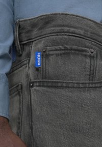 Dark gray jeans with a textured finish, featuring a blue "HUGO" tag, contrast stitching, and multiple pockets with black hardware.
