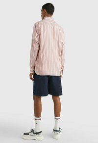 Camicia a maniche lunghe a righe rosa chiaro, pantaloni corti blu navy, calze bianche con bande rosse e nere, e scarpe nere con suole bianche.