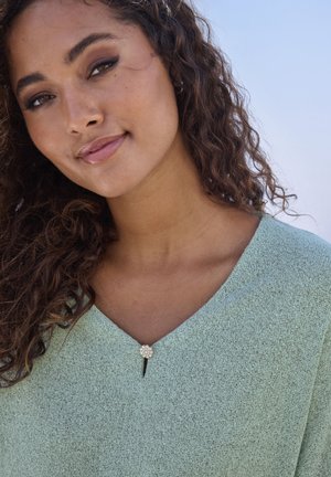 Woman with curly hair wearing a light green textured top with a pearl cluster brooch at the V-neckline against a clear sky background.