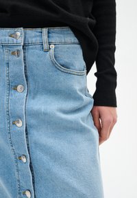 Jupe en denim bleu clair avec fermeture à boutons sur le devant, présentant une forme légèrement évasée, des poches visibles et des détails de surpiqûres contrastantes.
