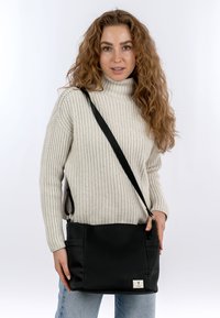 Schwarze gerippte Schultertasche mit flacher Form, ausgestattet mit einer Vordertasche und einem kleinen Logo-Patch. Das Modell trägt einen hellen Pullover und Jeans.