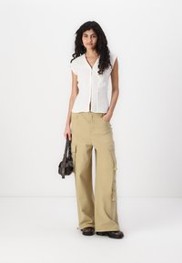 Chemisier blanc à manches courtes, associé à un pantalon cargo beige avec de grandes poches. La modèle tient un petit sac à main noir.