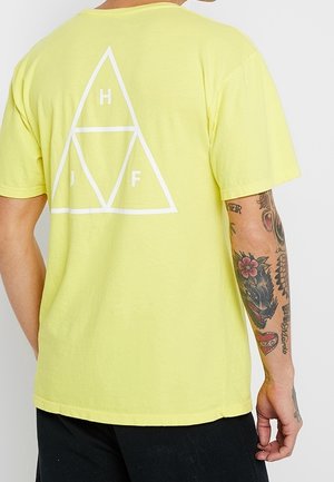 Personne portant un t-shirt jaune avec un motif de triangle blanc et les lettres H, J, F dans le dos, bras tatoué visible, pantalon noir.