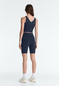 Woman standing facing away wearing navy blue sleeveless crop top and matching biker shorts with white sneakers and socks on white background.