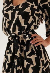 Abito nero e beige con motivo a fantasia e superficie texture, dotato di scollatura a V e dettaglio in vita con fiocco. Progettato con maniche a tre quarti.