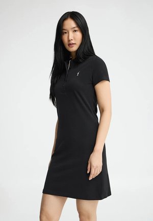 Kobieta z długimi czarnymi włosami ubrana w czarną sukienkę polo z krótkim rękawem i małym białym logo na piersi, stoi na tle jednolitego tła.