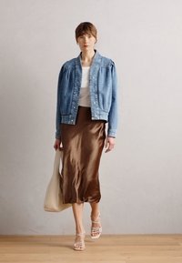 Traper jakna s nabranim ramenima, bijela majica i smeđa satenska midi suknja. Bež tote torba i sandale sa trakama upotpunjuju izgled.