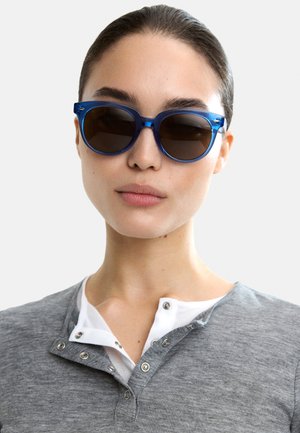 Niebieskie plastikowe okulary przeciwsłoneczne z okrągłymi oprawkami, ciemnymi soczewkami i błyszczącym wykończeniem. Noszone z szarym T-shirtem henley z zatrzaskami.