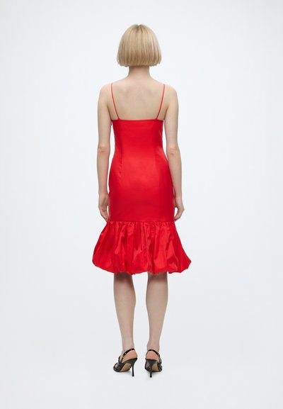 Femme aux cheveux blonds courts portant une robe en satin rouge cintrée avec de fines bretelles et un ourlet volanté, associée à des sandales noires à talons hauts.