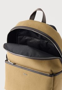 Καφέ σακίδιο από ύφασμα με μαύρο εσωτερικό, μπροστινή τσέπη με φερμουάρ, επάνω λαβή και ασημένιο λογότυπο BOSS στη μπροστινή τσέπη.