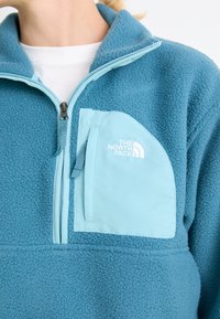Fleece pullover in teal with a quarter-zip collar. Features a light blue chest pocket with a zip closure and subtle branding. Soft texture.