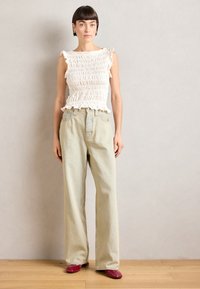 Hvit smocket topp med volanger, kombinert med lyseblå vide jeans. Modellen har på seg røde spisse sko. Nøytral bakgrunn.