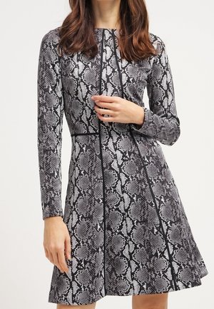 Vrouw draagt een lange mouw, knielange jurk met een zwart en grijs slangenhuidpatroon en verticale zwarte naaddetails.