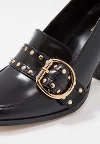 Chaussure en cuir noir avec une large sangle ornée d'une boucle dorée et de clous décoratifs. Le talon est de hauteur modérée, apportant une certaine structure.