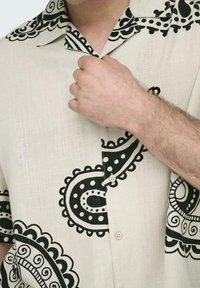 Kurzarmhemd aus hellbeigem Stoff, mit schwarzen Paisley-Mustern, spitzem Kragen und Knopfverschluss vorne.