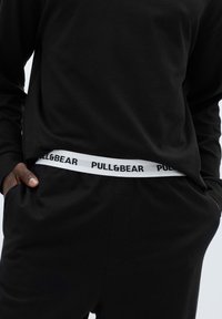 Černá mikina a kalhoty mají bílý elastický pás s nápisem „PULL&BEAR“ v černé barvě. Materiál působí hladce a pohodlně.
