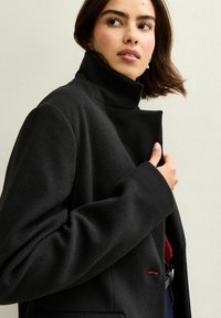 Svart ullblandad kappa med hög krage, strukturerade axlar och dold knapplist; har en röd accent på fickan.