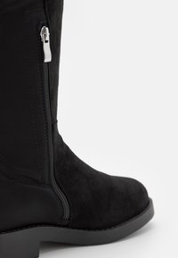 Botín de ante negro con cremallera lateral, tacón de goma bajo y acabados suaves. Presenta un diseño estructurado y costuras mínimas.