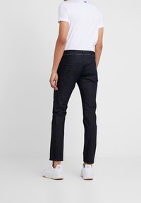 Jean en denim foncé à coupe slim, avec cinq poches, des coutures visibles et un logo de marque discret sur la ceinture arrière. Porté avec un t-shirt blanc.