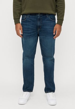 Dunkelblaue Jeans mit geradem Bein, subtilen Verwaschungen und einem Fünf-Taschen-Design. Kombiniert mit weißen Sneakers und einem grünen Hoodie.