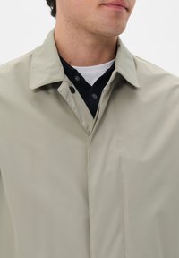 Chaqueta impermeable de color gris claro con un cuello con solapa y costuras mínimas. Presenta una tapeta abotonada y una textura suave y lisa.