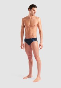 Uomo in forma che sta in piedi a piedi nudi, indossando slip da bagno scuri con accenti blu, guardando verso sinistra su uno sfondo grigio chiaro uniforme.