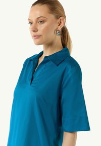 Türkise Hemd mit Kragen, V-Ausschnitt und kurzen Ärmeln. Glatte Textur, lässiges Design mit einer dezenten seitlichen Nahttasche.