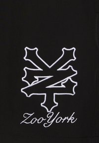 Black fabric featuring a white embroidered logo resembling a stylized letter 'Z' with sharp accents, accompanied by the text "Zoo York" below.