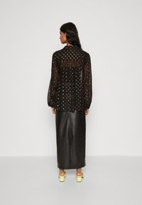 Schwarze halbtransparente Bluse mit mehrfarbigem Punktenmuster, langen Ärmeln und hohem Kragen. Kombiniert mit einem langen schwarzen Lederrock und gelben Schuhen.