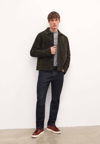 Chaqueta de ante verde oscuro con botones frontales, camiseta gris debajo, jeans oscuros y zapatillas de cuero marrón sobre un fondo neutro.