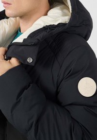 Zwarte pufferjack met een witte capuchonvoering, voorzien van een ritssluiting en een rond beige logo-opdruk op de linkermouw. Geverfde stof, geïsoleerd.