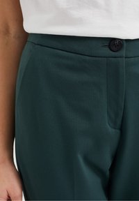 Nahaufnahme von dunkelgrünen, taillierten Hosen mit einem schwarzen Knopf und seitlichen Taschen, getragen mit einem in die Hose gesteckten weißen Oberteil von einer Person.